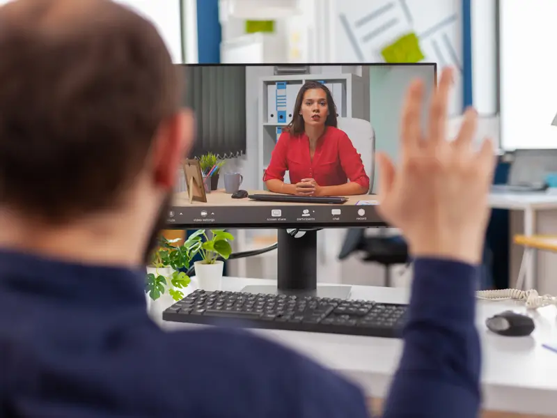 A person seen from behind waves at a desktop monitor displaying a video call with a woman in a red blouse seated at a professional desk. The setup illustrates a remote job interview or executive assistant screening conducted over video conferencing software.