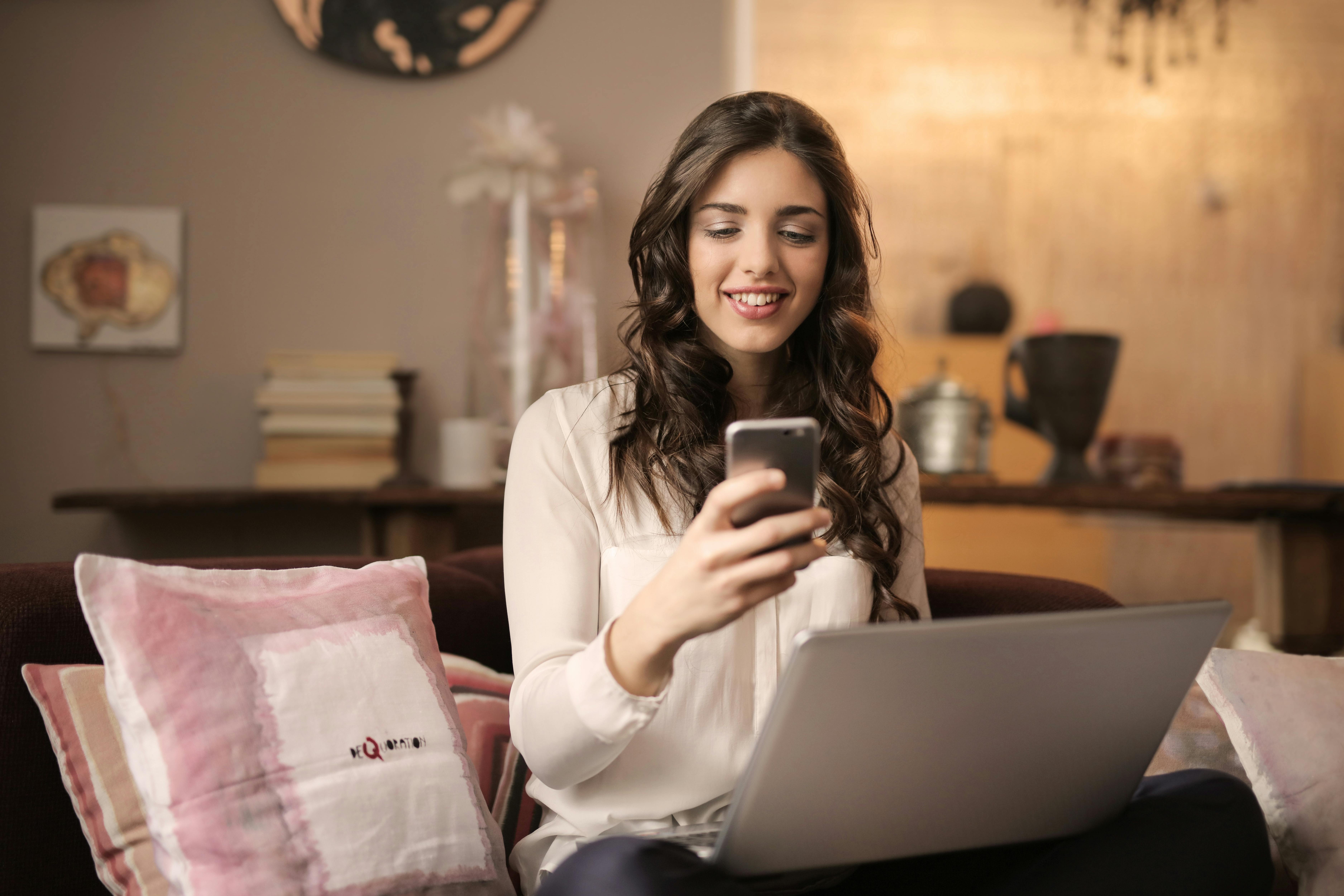 Smiling woman sitting on a sofa with a laptop, checking her phone while researching finance staffing agencies
