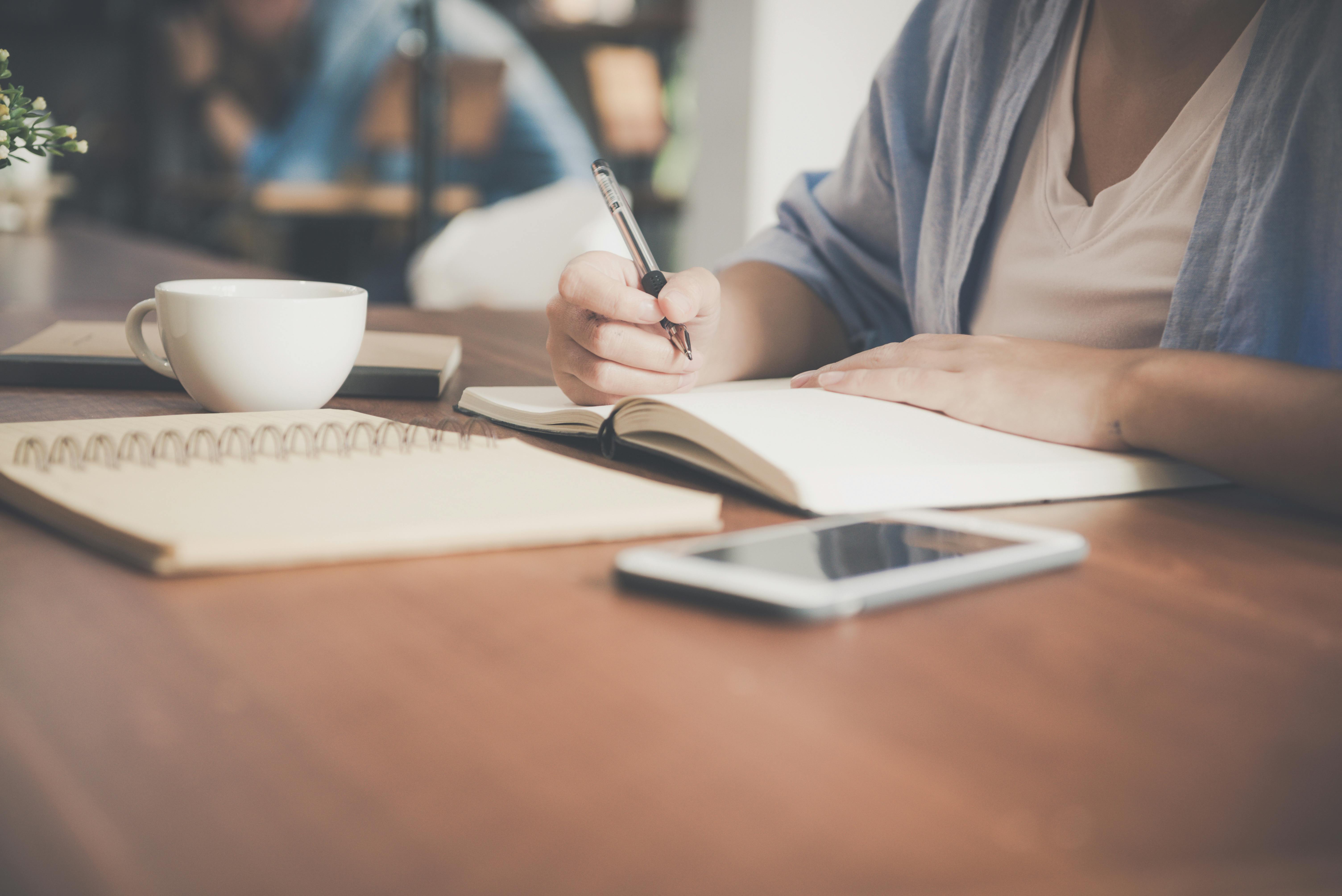 Person writing notes in a notebook at a wooden desk with a coffee cup and smartphone nearby