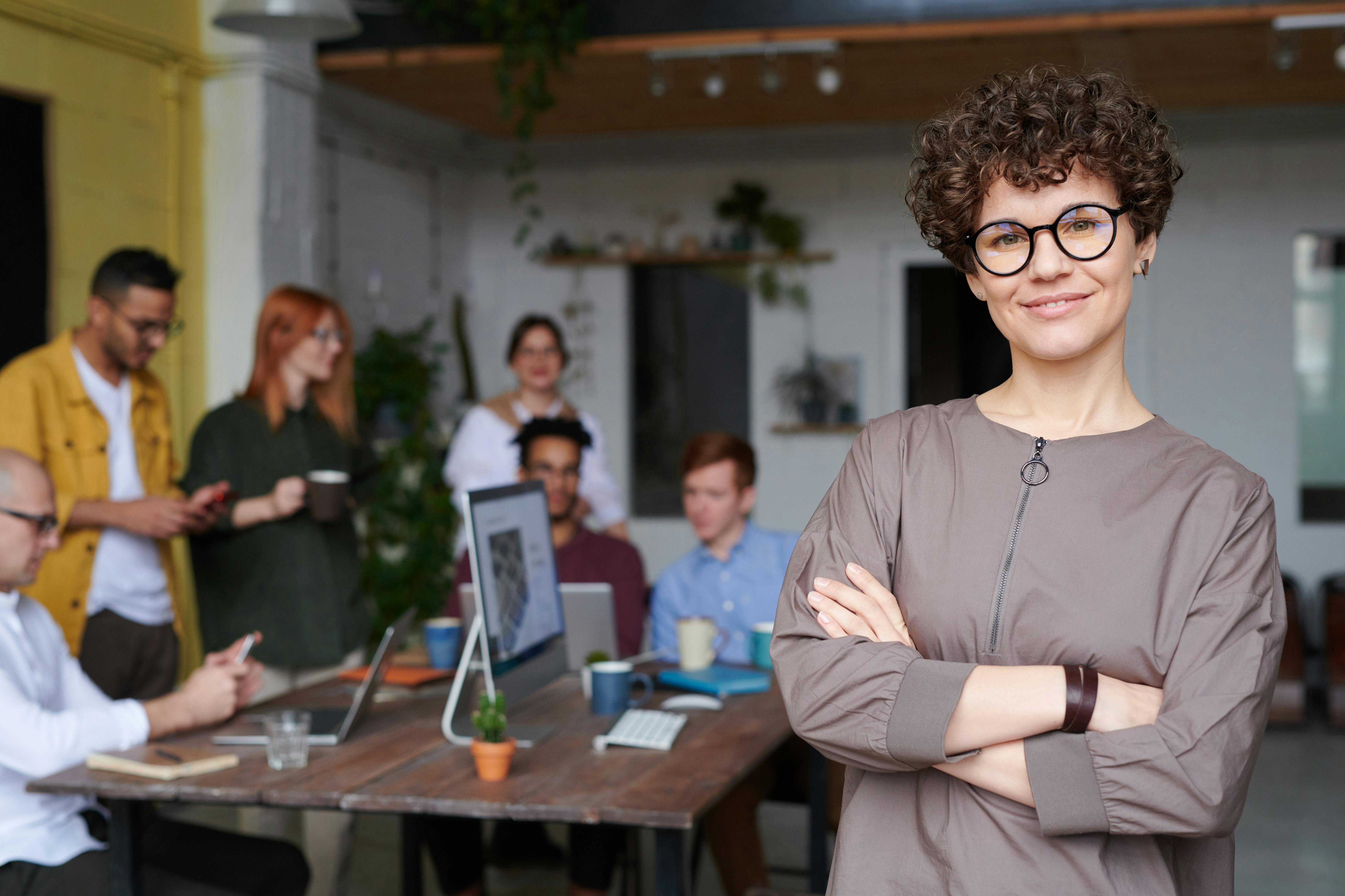 A confident woman with short curly hair and glasses stands with arms crossed in a modern open-plan office, smiling at the camera. In the background, a diverse team of colleagues works at a shared table with laptops, suggesting a project management or team leadership context.