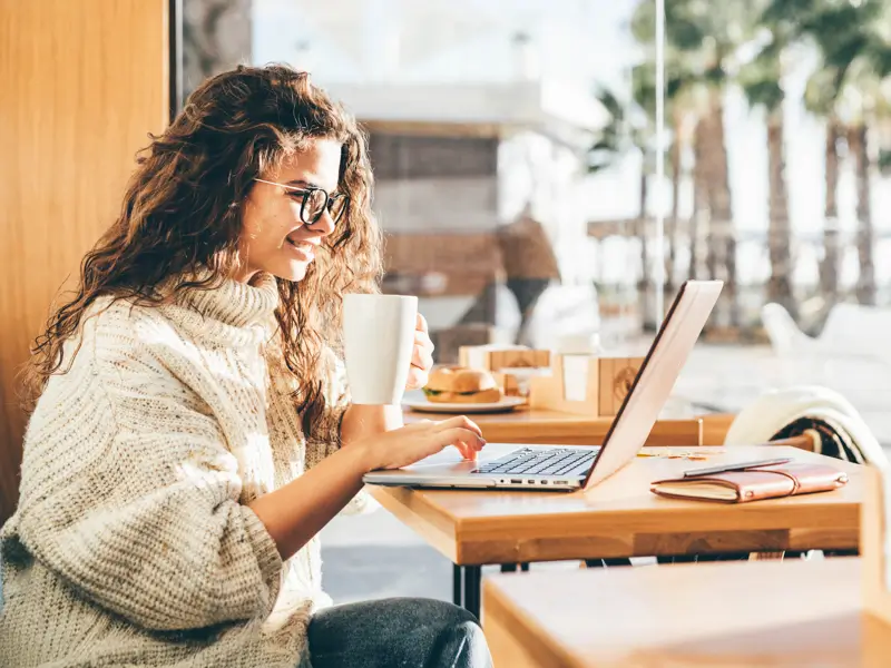 A smiling young woman with curly hair and glasses holds a white coffee mug while working on a laptop at a wooden café table. Food and a notebook are visible nearby, with palm trees and sunlight visible through the window behind her, evoking a relaxed remote work setting.