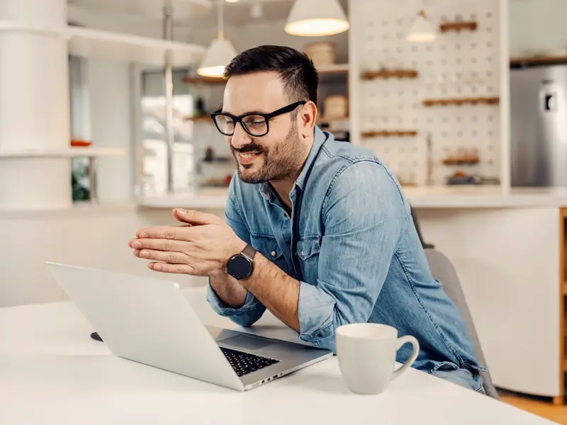 Man with glasses smiling and gesturing during a video call on a laptop in a bright kitchen workspace