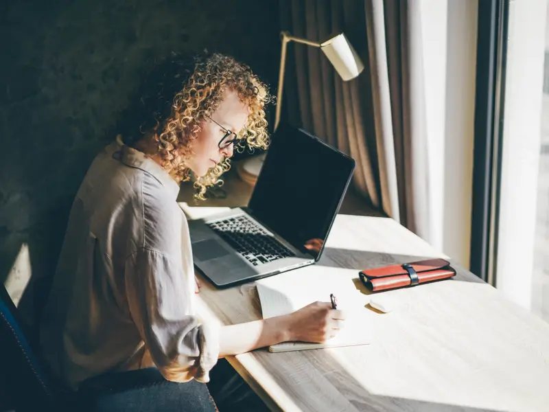 Woman with curly hair and glasses writing notes by hand beside a laptop at a desk near a window