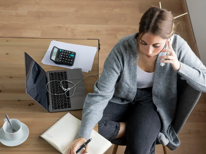 Overhead view of a woman in a grey cardigan sitting on a chair, talking on a smartphone while writing in a notebook. A laptop, calculator, printed documents, wired earphones, and a coffee cup are arranged on a wooden floor around her, suggesting a remote accounts receivable or finance work session.
