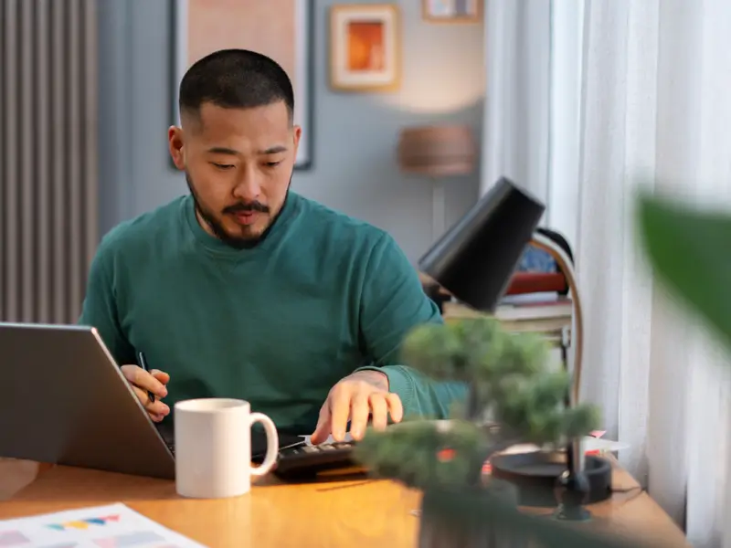 An Asian man in a teal sweater sits at a home office desk, using a calculator while working on a laptop. A white mug, small potted pine tree, desk lamp, and financial documents with colorful charts are visible on the desk, conveying focused accounts receivable or bookkeeping work.