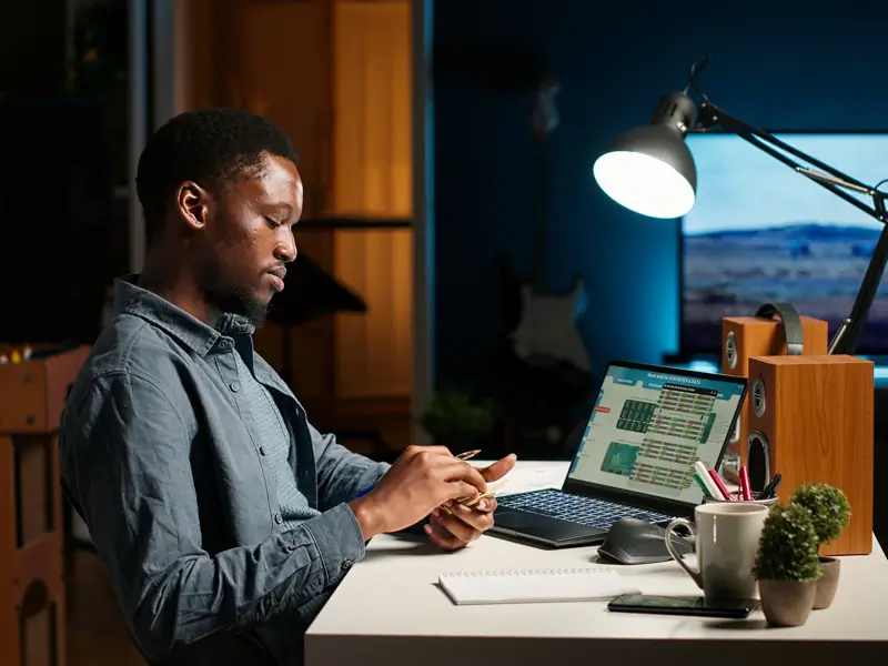 A Black man in a denim shirt works at a desk under a bright lamp, reviewing a financial spreadsheet on a laptop screen with color-coded rows and columns. A notebook, mug, small plant, and audio speakers are visible in the background of what appears to be a home office set up for late-night financial work.
