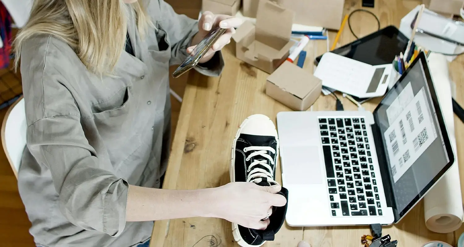 Woman photographing a sneaker with her phone at a desk covered in cardboard shipping boxes, with a laptop showing product listings beside her