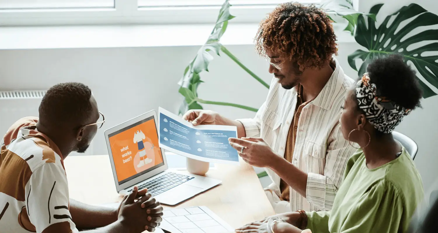 Three colleagues at a table reviewing a printed infographic and a laptop showing a "What works best?" slide during a team discussion