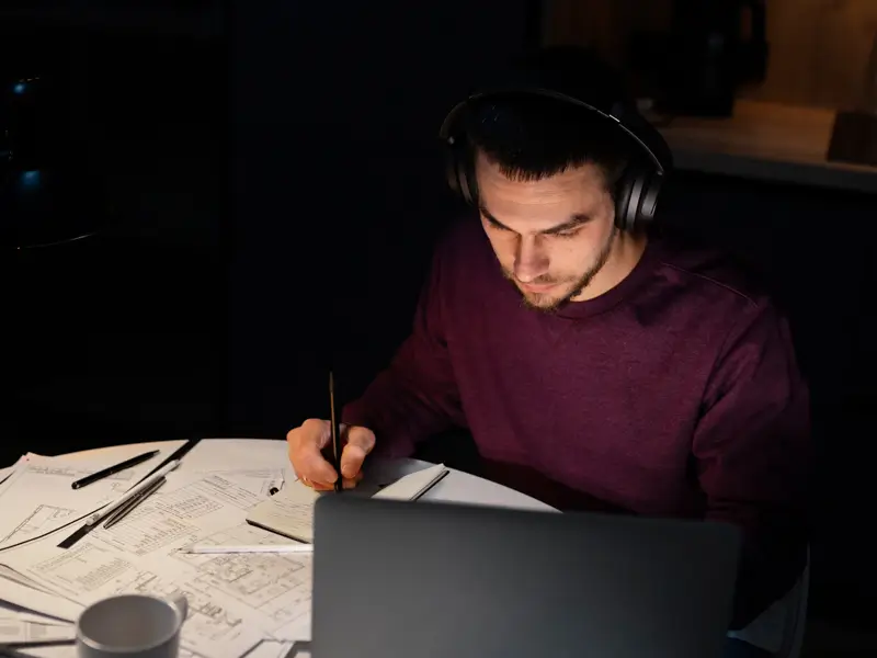 A man in a dark maroon sweater and headphones sits at a desk late at night, writing notes by hand on technical documents or diagrams spread across the surface, with a laptop open nearby. The scene is dimly lit, suggesting intensive review or QA documentation work.