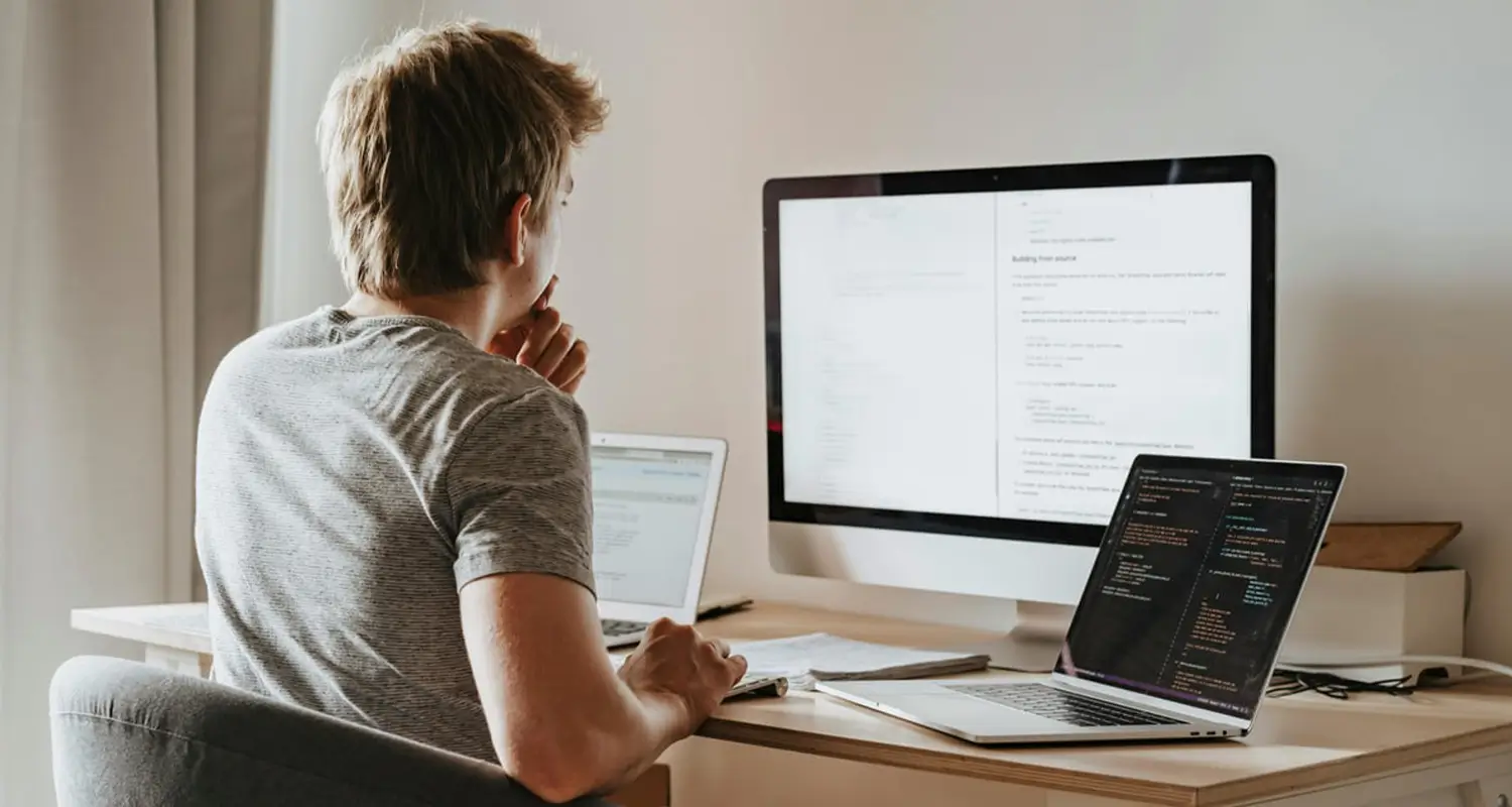 A remote developer seen from behind, working at a home desk with a large desktop monitor displaying a document with code, a dark-themed laptop showing colorful syntax-highlighted code, and a tablet, illustrating a multi-screen remote software development setup.