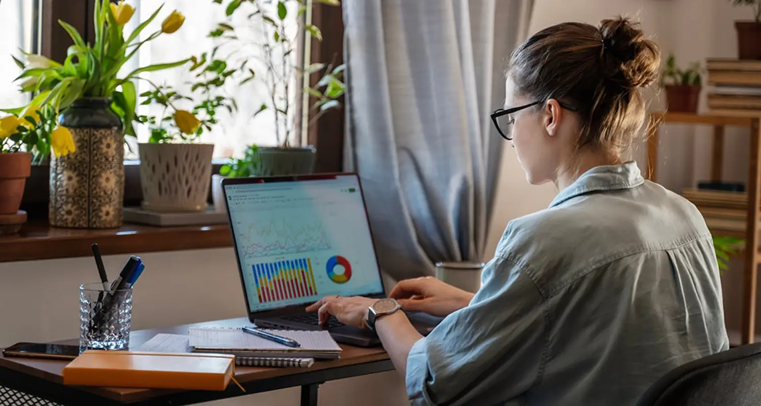 A woman with glasses and her hair up works from a home office surrounded by potted plants and yellow tulips, reviewing colorful bar charts and a pie chart on her laptop screen alongside handwritten notes, representing a remote analyst or data professional in a comfortable home workspace.