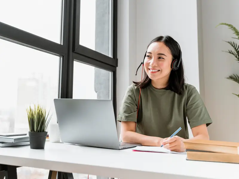 A smiling woman wearing a headset with a microphone sits at a bright, modern desk with a laptop, notebook, and small plant. She holds a pen and appears to be taking notes, representing a virtual assistant working remotely in a well-lit workspace with large windows.