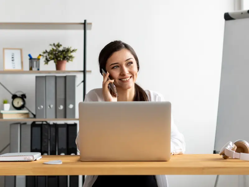 A young woman in a white blouse smiles while talking on a smartphone at a tidy office desk with a laptop, notepad, and wireless headphones. Shelves with binders, a plant, and a clock are visible in the background, suggesting a professional virtual assistant handling client communications.
