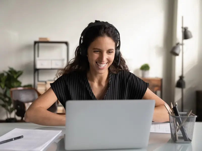 A smiling woman with curly hair wears over-ear headphones and sits at a bright home office desk with a laptop open in front of her. A notepad, pen, and desk organizer are visible nearby, depicting a cheerful virtual assistant engaged in a remote work session.