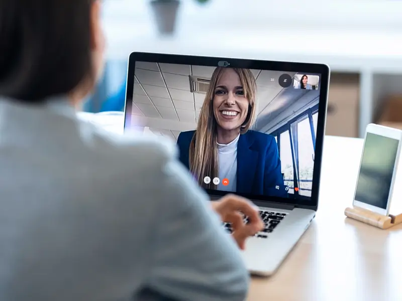Viewed from behind, a person in a grey top sits at a desk with a laptop open to a video call showing a smiling woman in a blue blazer and white top. A small self-view thumbnail is visible in the top corner of the screen, and a smartphone in a stand sits nearby, depicting a professional remote job interview or hiring conversation.