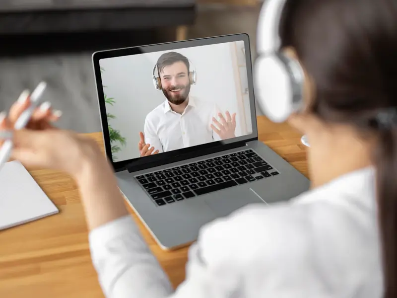 Viewed from behind, a woman with dark hair in a ponytail and white over-ear headphones sits at a wooden desk, gesturing with one hand while conducting a video interview. The laptop screen shows a smiling bearded man in a white shirt and headset waving, illustrating a remote job interview in progress between a recruiter and a candidate.
