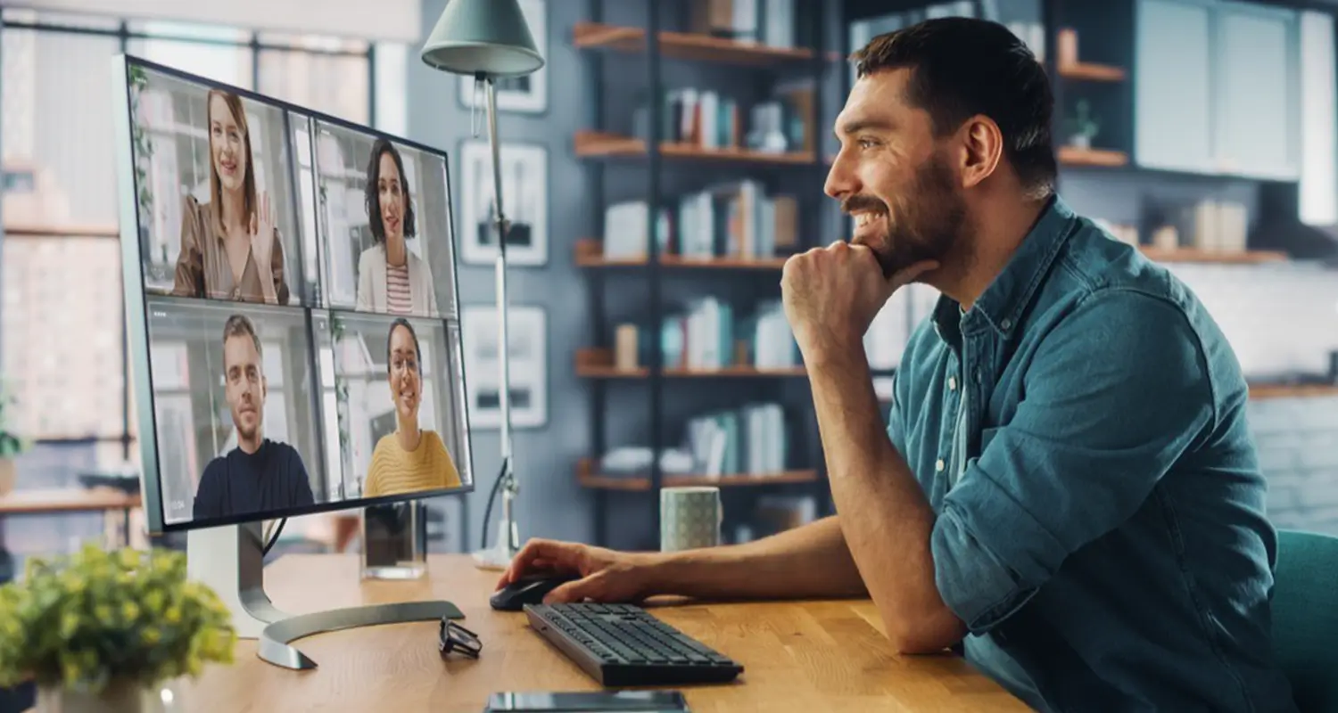 Man smiling during a video call with four colleagues shown in a grid on a large desktop monitor at his home office.