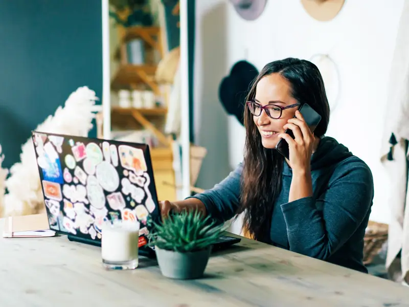 A smiling woman wearing glasses talks on her phone while working on a sticker-covered laptop at a home workspace. A small potted succulent and a candle sit on the wooden table in front of her, representing a remote worker or recruiter managing communications from a casual home office environment.