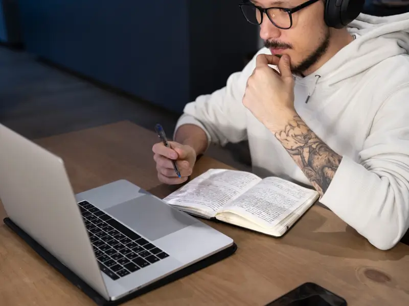 A young man with tattoos, wearing a white hoodie and over-ear headphones, studies intently at a dark desk. He holds a pen over an open notebook while looking at a laptop screen, depicting focused research or content work typical of an SEO professional or remote knowledge worker.
