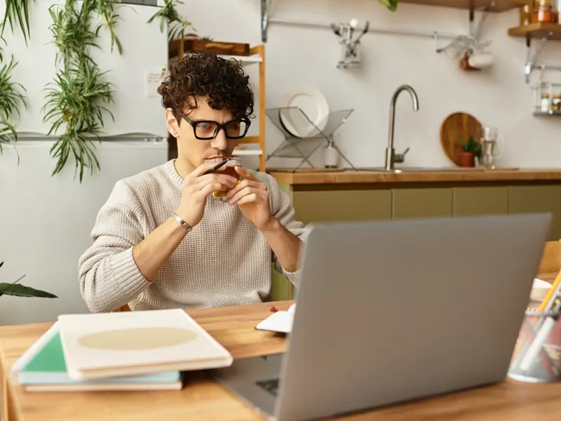 A young man with curly hair and glasses sits at a wooden desk in a home kitchen setting, holding a small glass while reviewing content on an open laptop. Notebooks and stationery are visible on the desk, suggesting focused remote work or candidate research.