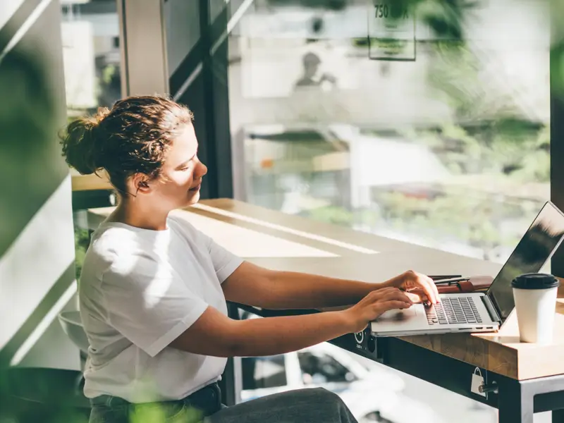 A woman with her hair up in a bun sits at a wooden table near a large sunlit window in a cafe-like setting, actively typing on a laptop. A takeaway coffee cup sits beside her, conveying a productive remote or hybrid work session.