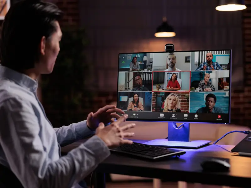 Viewed from behind, a person in a light blue shirt sits at a desk in a warmly lit room, gesturing with both hands during a video conference. The monitor shows a nine-person grid of remote participants, representing a virtual team meeting with augmented or distributed staff members.