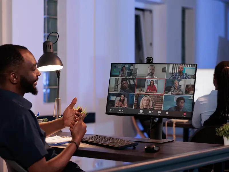 A Black man with a beard sits at a desk in a dimly lit office, gesturing with his hands while participating in a video call. A large monitor displays a grid of nine remote participants from diverse backgrounds in what appears to be a team meeting, illustrating a distributed workforce collaborating virtually.