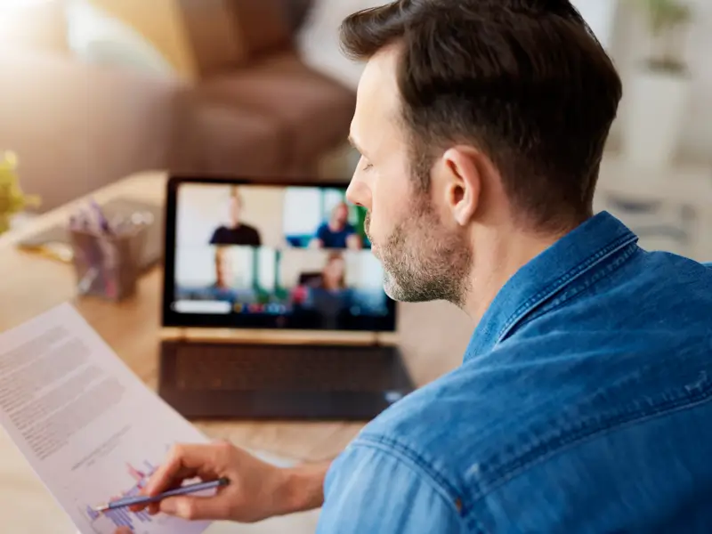 A man with dark hair and a short beard, wearing a denim shirt, holds printed documents with charts while looking at a laptop screen showing a small video call with colleagues. He appears to be working from home, reviewing materials during a remote team collaboration session.