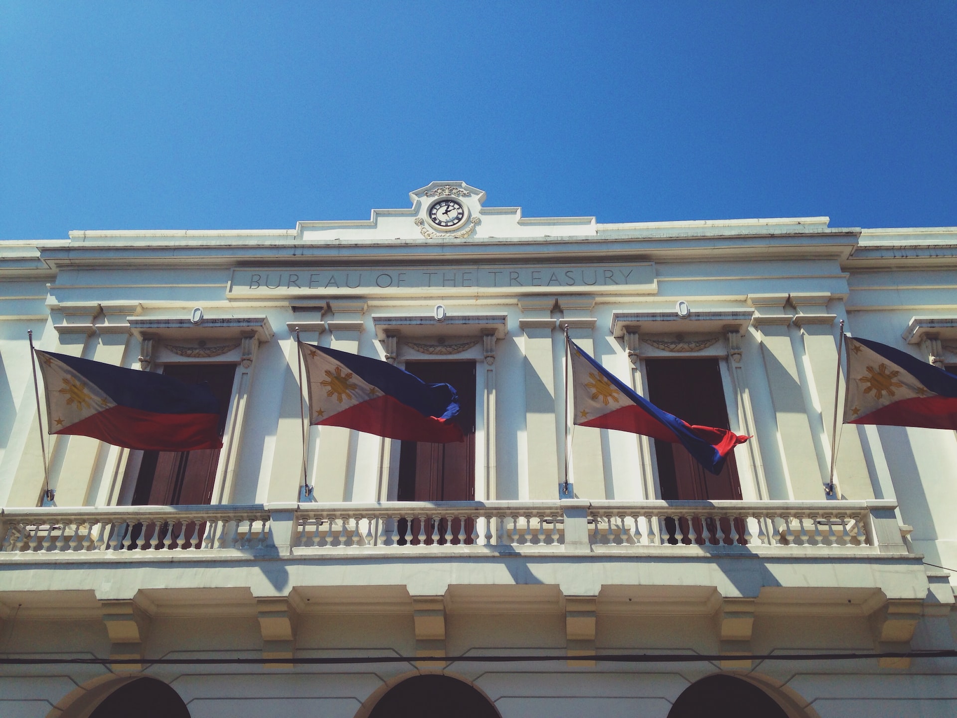 The facade of the Philippines Bureau of the Treasury building, a white colonial-style structure with a clock at the top and four Philippine flags displayed across its balcony under a clear blue sky.