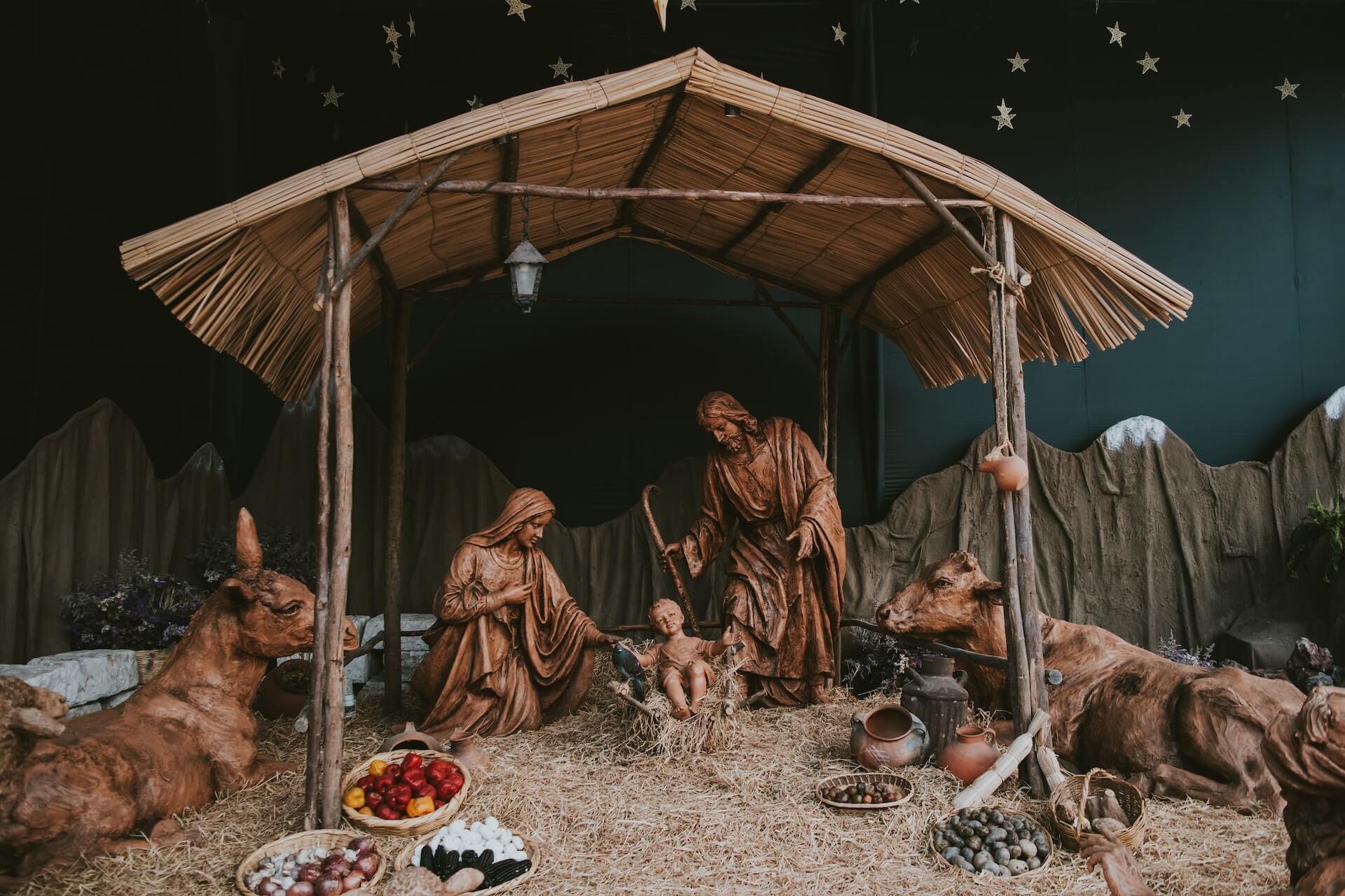 A detailed nativity scene display with carved wooden figures of Mary, Joseph, and the infant Jesus in a manger, surrounded by animals including a donkey and cattle. Star decorations hang overhead and bowls of food and items are arranged at the base, reflecting the Philippines' strong Christmas tradition.