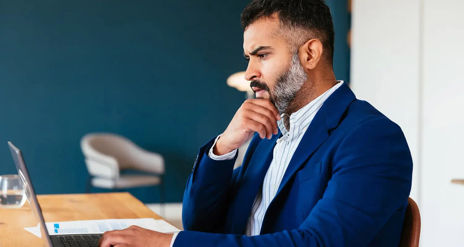 Focused professional man with beard in a blue blazer working on a laptop at a wooden desk in a modern home office