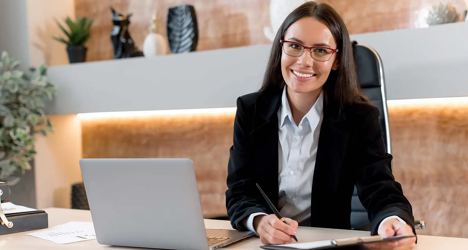 Smiling professional woman with glasses and dark hair sitting at a desk with a laptop and notepad in a warmly lit office