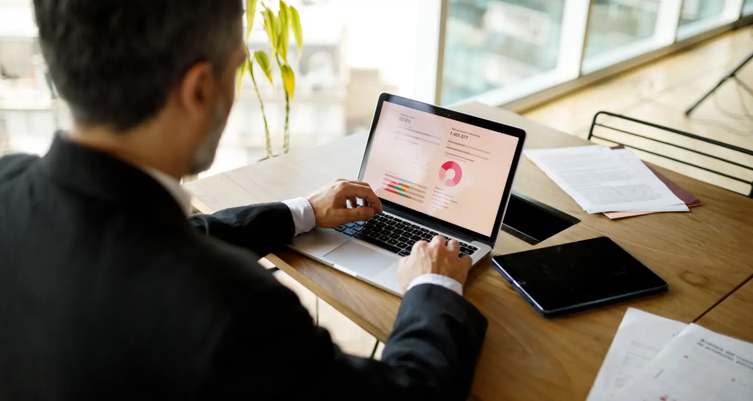 Businessman from behind working on a laptop displaying a colorful analytics dashboard with pie charts and bar graphs in a bright office