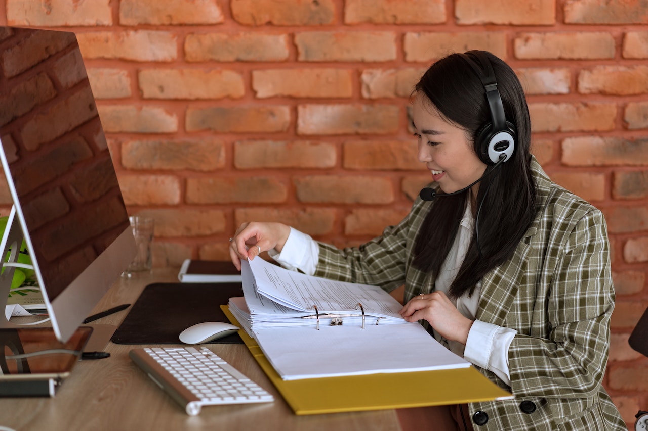 A smiling Asian woman wearing a headset sits at a desk with a desktop computer, keyboard, and mouse, flipping through pages in a large binder. She appears engaged and professional, representing a data entry or administrative specialist working remotely.