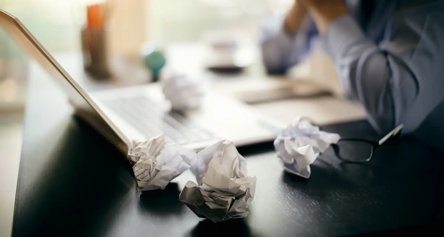 Close-up of crumpled paper balls scattered across a dark desk surface, with an open laptop and a blurred figure of a person holding their head in the background, symbolizing repeated failed attempts and workplace frustration.
