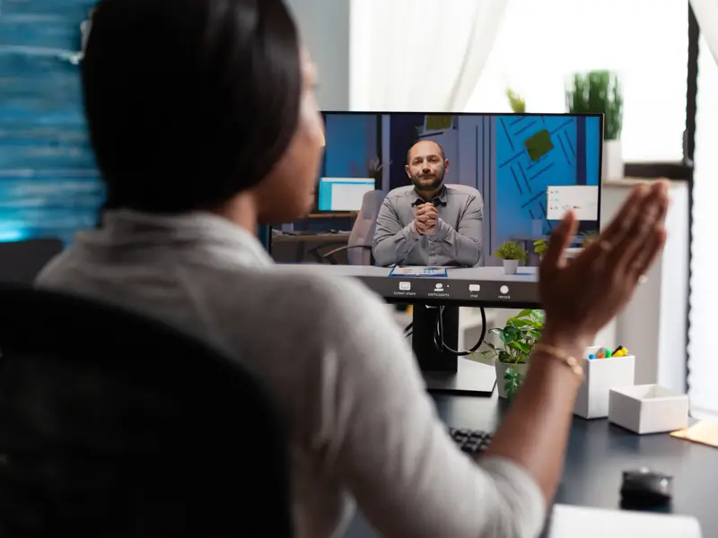 A woman seen from behind gestures with her hand while conducting a remote video interview at a home office desk. On the monitor in front of her, a man in a grey shirt sits with hands clasped in a professional setting. Office supplies and plants are visible on the desk in the foreground.