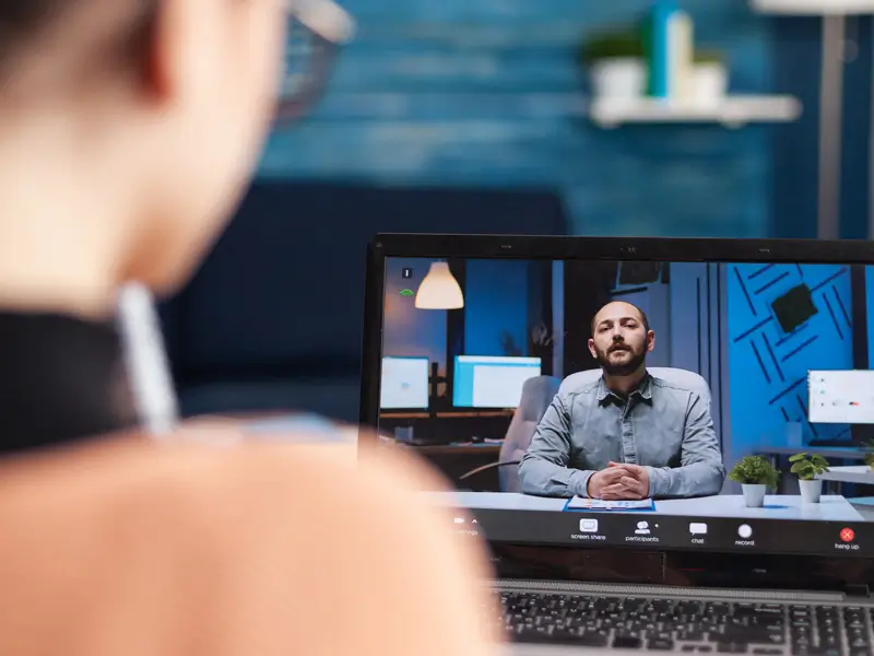 A woman seen from behind looks at a laptop screen during a video interview. On the screen, a man in a denim shirt sits with hands clasped at a desk in a softly lit home office. Video call controls including screen share, participants, chat, and record are visible at the bottom of the screen.