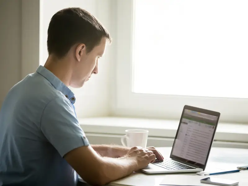 Man in a blue shirt focused on a laptop by a bright window with a coffee mug beside him