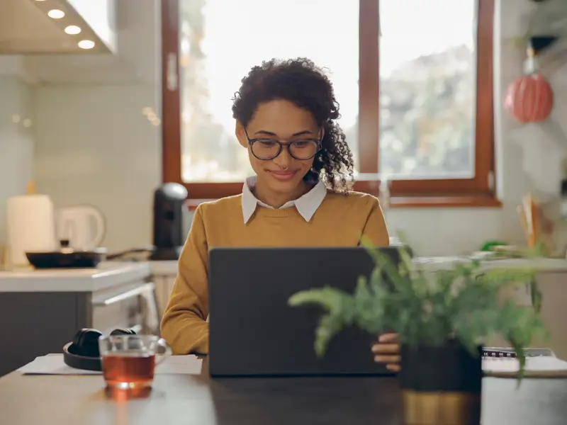 Woman with curly hair and glasses smiling while working on a laptop at a home kitchen table with tea and a plant