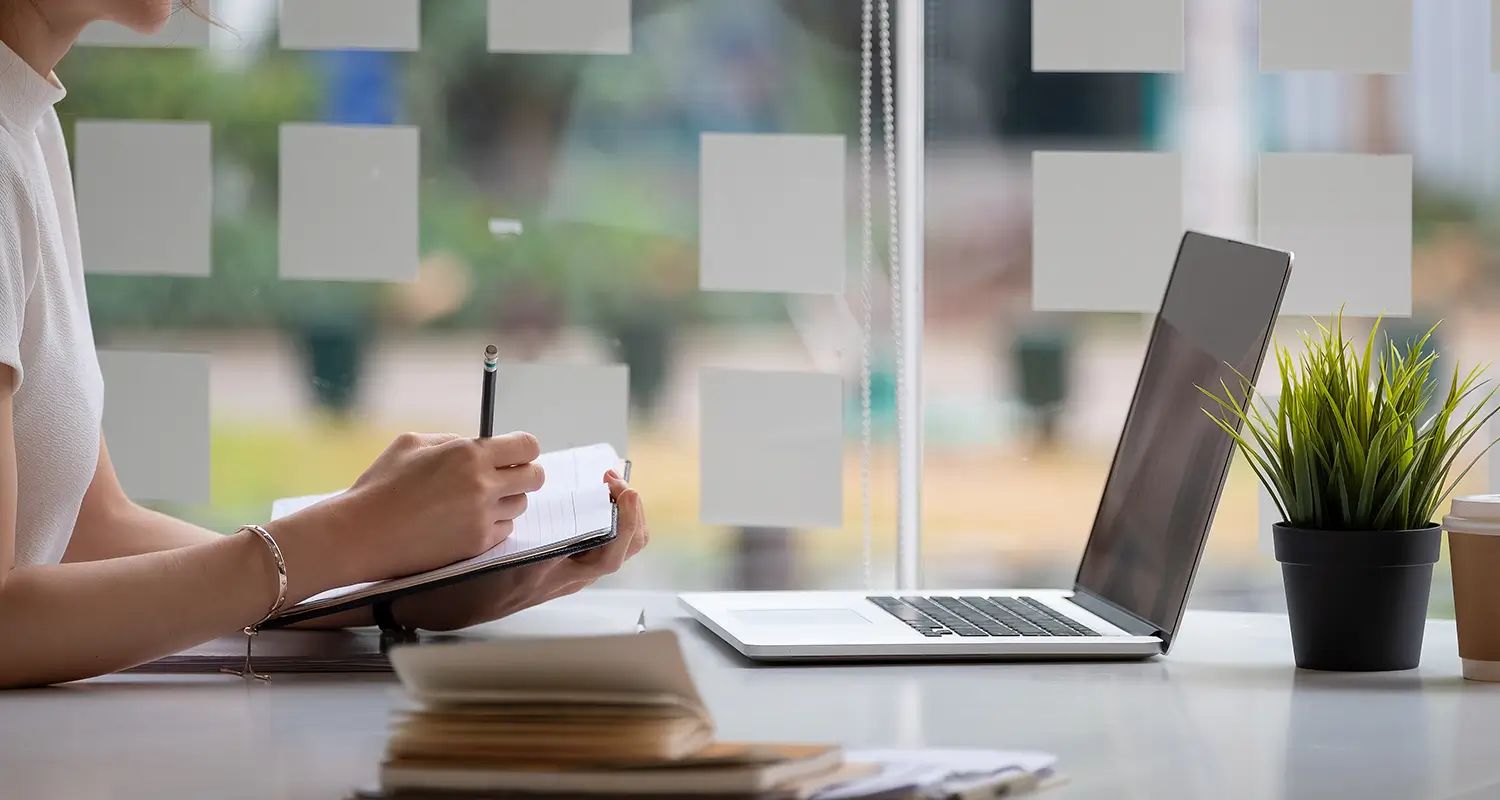 Person writing notes in a notebook beside a laptop at a desk with sticky notes on a glass window, planning and organizing work tasks.