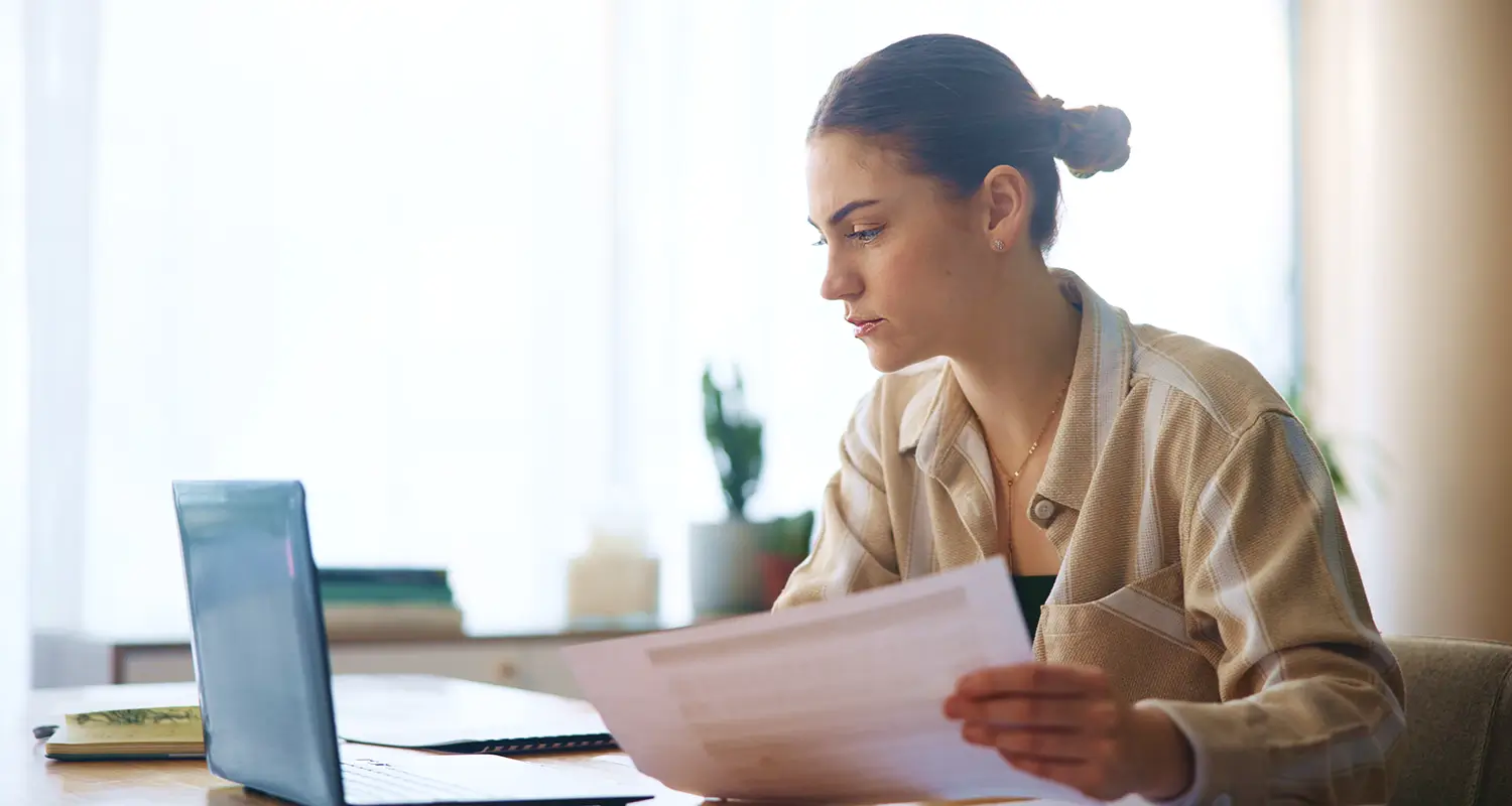 Woman reviewing printed documents at a desk with a laptop, focused on evaluating written materials in a home office setting.