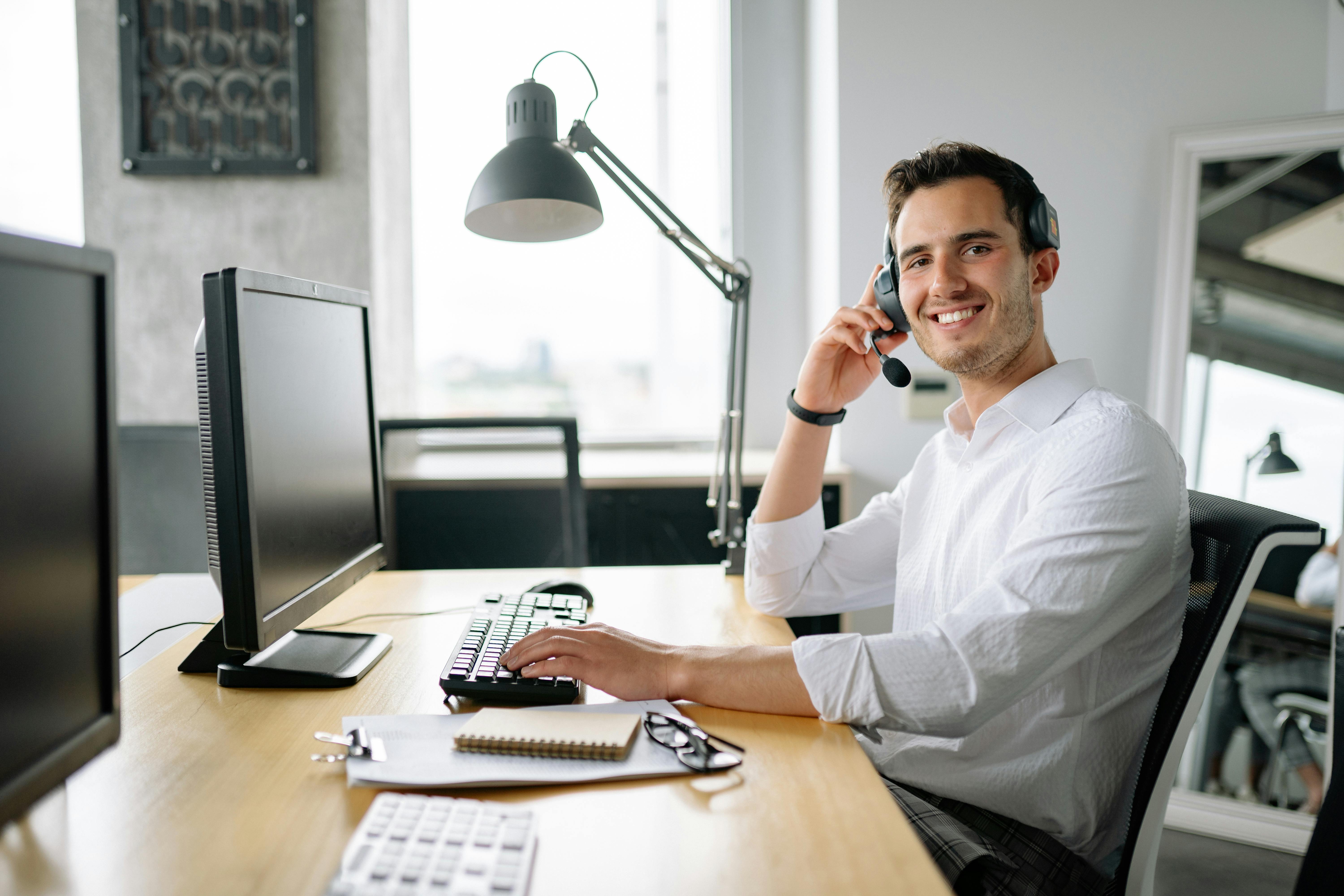 A smiling man in a white shirt wearing a headset sits at an office desk with a desktop computer and keyboard, looking toward the camera. He represents a customer support or recruitment specialist working at a South African staffing firm.