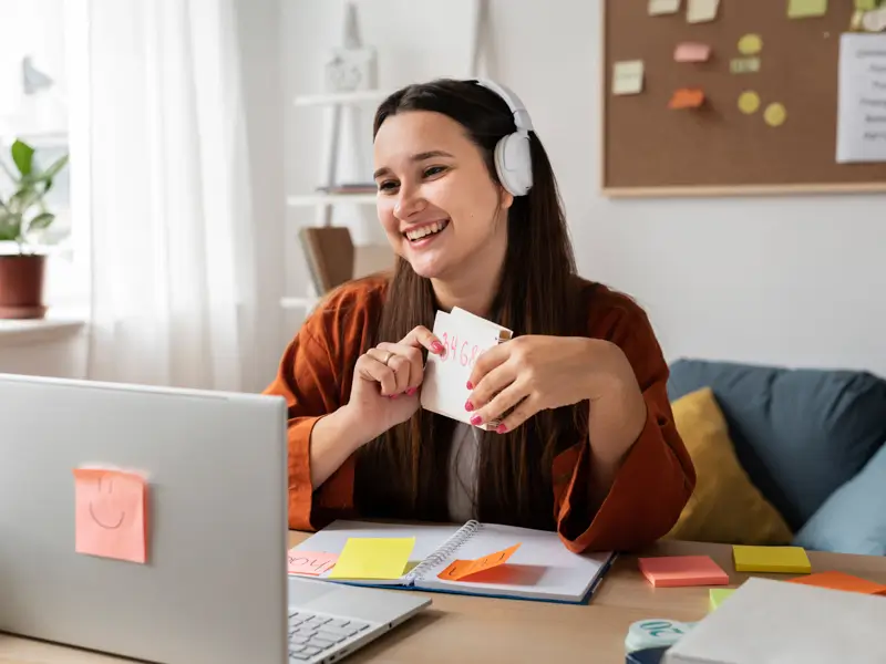 Woman with headphones smiling and holding a sticky note at a desk covered with colorful sticky notes and a notebook