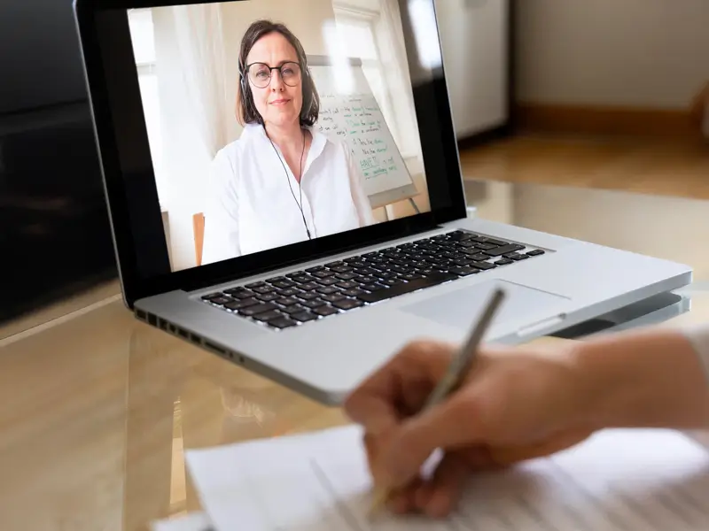 Person taking notes by hand while a woman with headset and glasses appears on a laptop screen during a video call