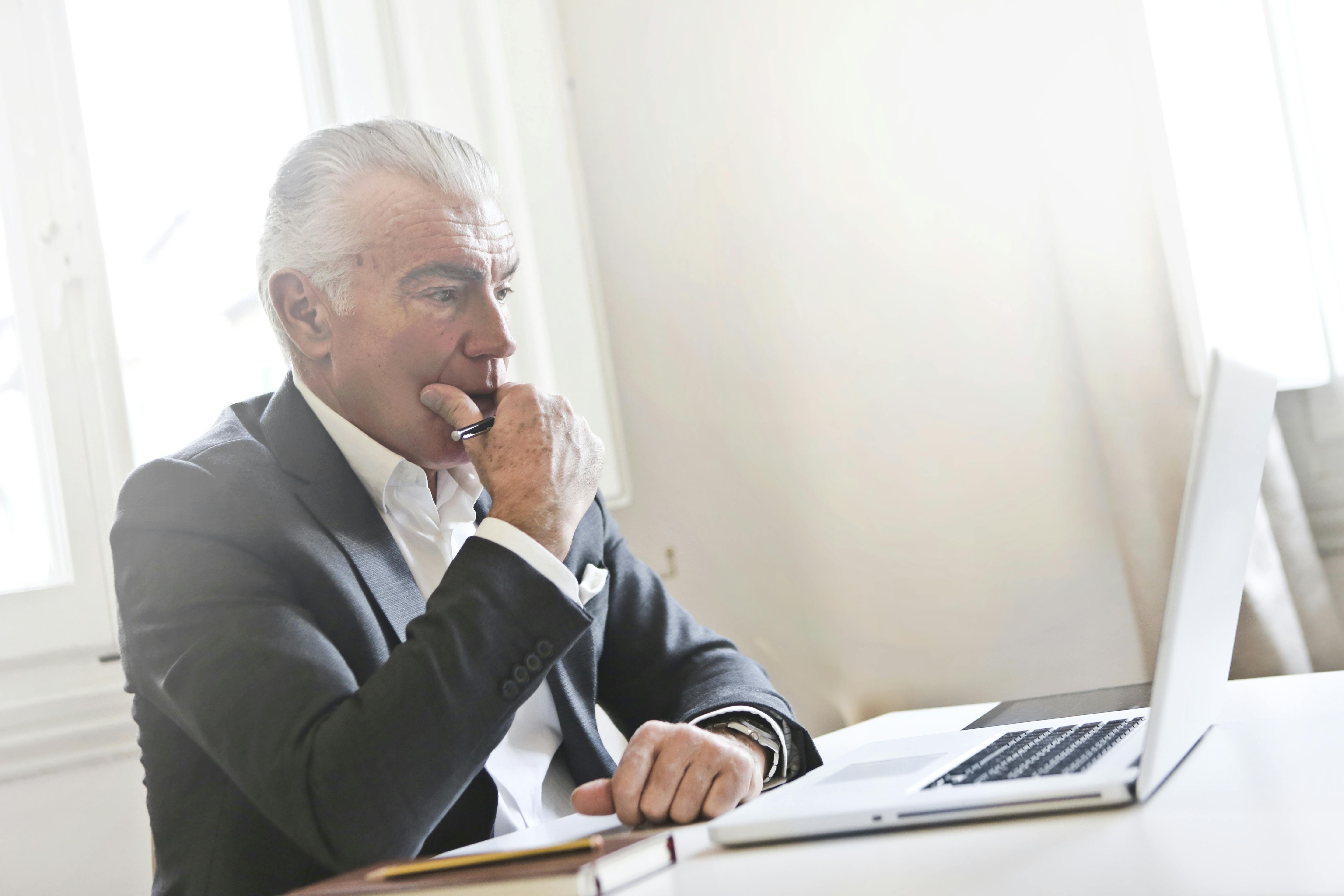 Senior executive man in a dark suit sitting at a desk, pensively reviewing content on a laptop