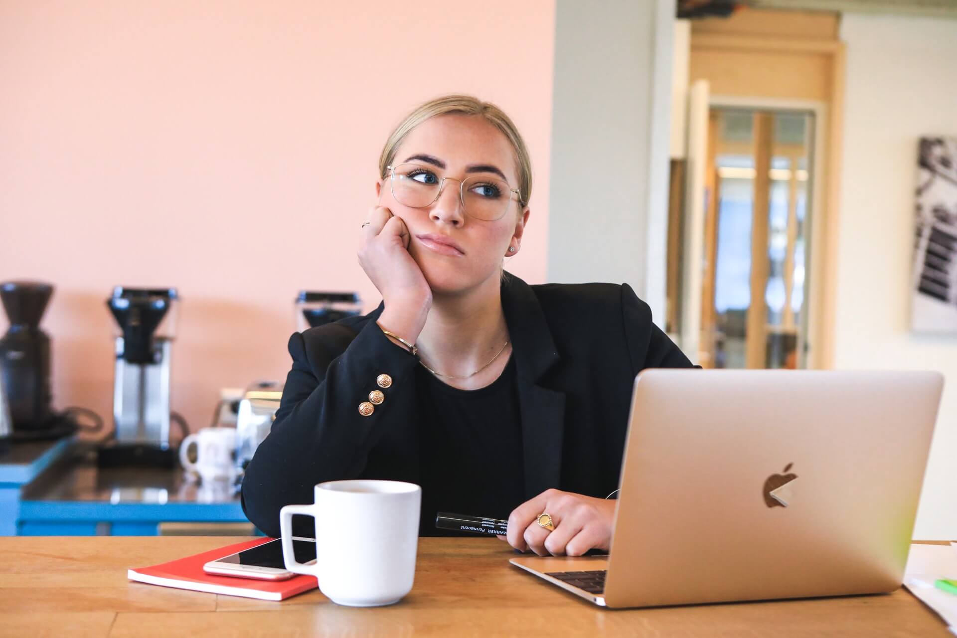 A young woman with blonde hair and round glasses wearing a black blazer rests her chin on her hand with a bored or frustrated expression while sitting in front of an open MacBook laptop and a white coffee mug. The image depicts an overwhelmed business owner who lacks a clear onboarding process for new virtual assistants.