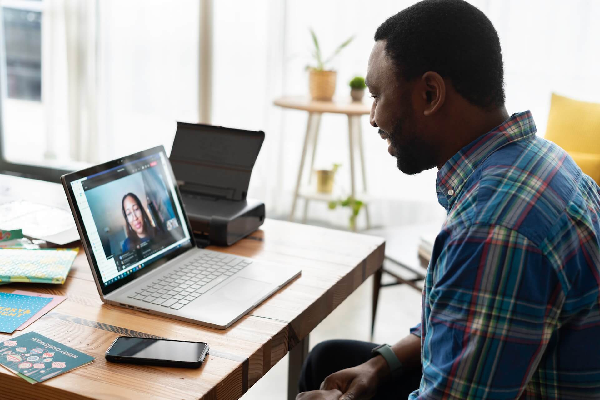 A man in a plaid shirt sits at a wooden home office desk and conducts a video call on a laptop showing a woman on screen, with a smartphone resting on the desk beside the computer. The scene represents a remote employer meeting with a new virtual assistant during the onboarding introduction stage.