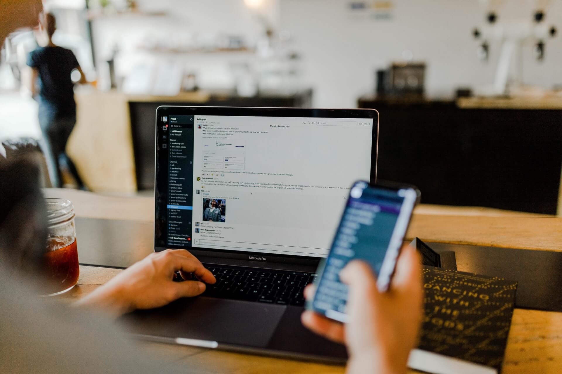 A person at a café table uses a MacBook Pro displaying a Slack workspace with active message threads, while holding a smartphone showing the Slack mobile app. The scene illustrates using team communication tools during the virtual assistant onboarding process to stay connected with a remote employer.