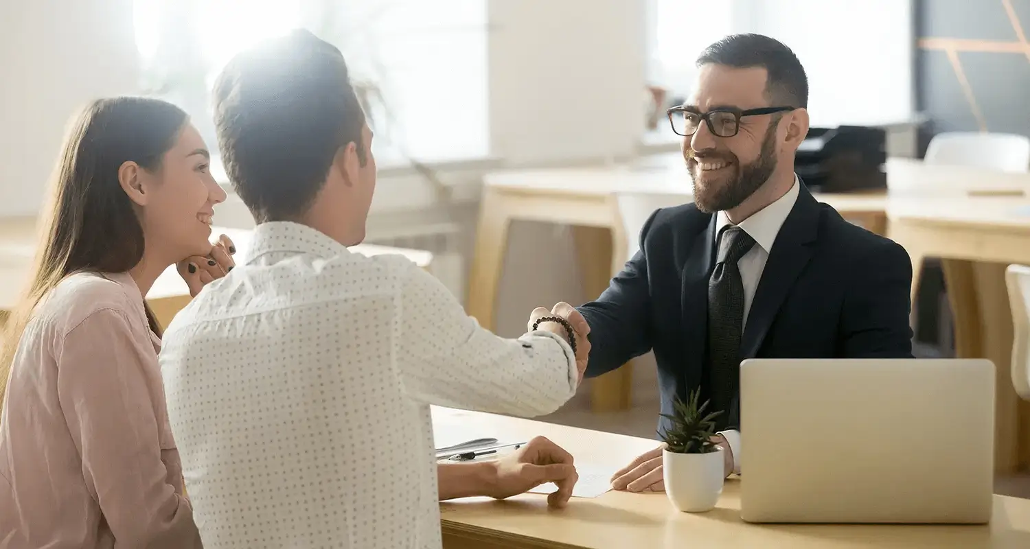 A bearded property manager in a dark suit and tie shakes hands with a male client across a desk while the client's female partner smiles beside him. A laptop and small potted plant sit on the desk in a bright office, depicting a successful property management agreement being finalized.