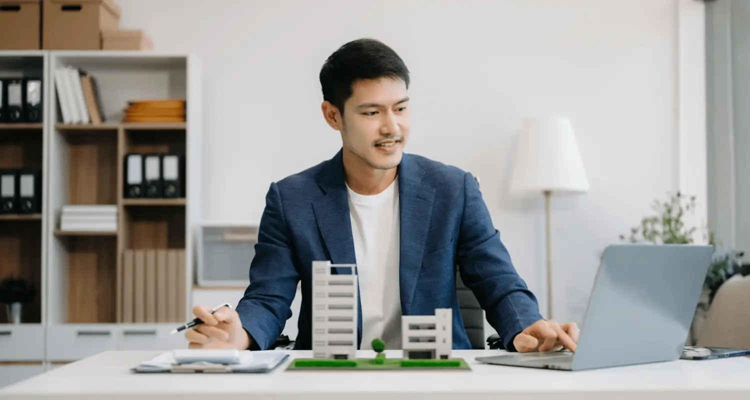 A man in a blue blazer sits at a clean office desk reviewing content on a laptop, with a pen in hand and two small architectural building models placed in front of him. Bookshelves with binders and a lamp are visible in the background, suggesting a property portfolio management or compliance review session.